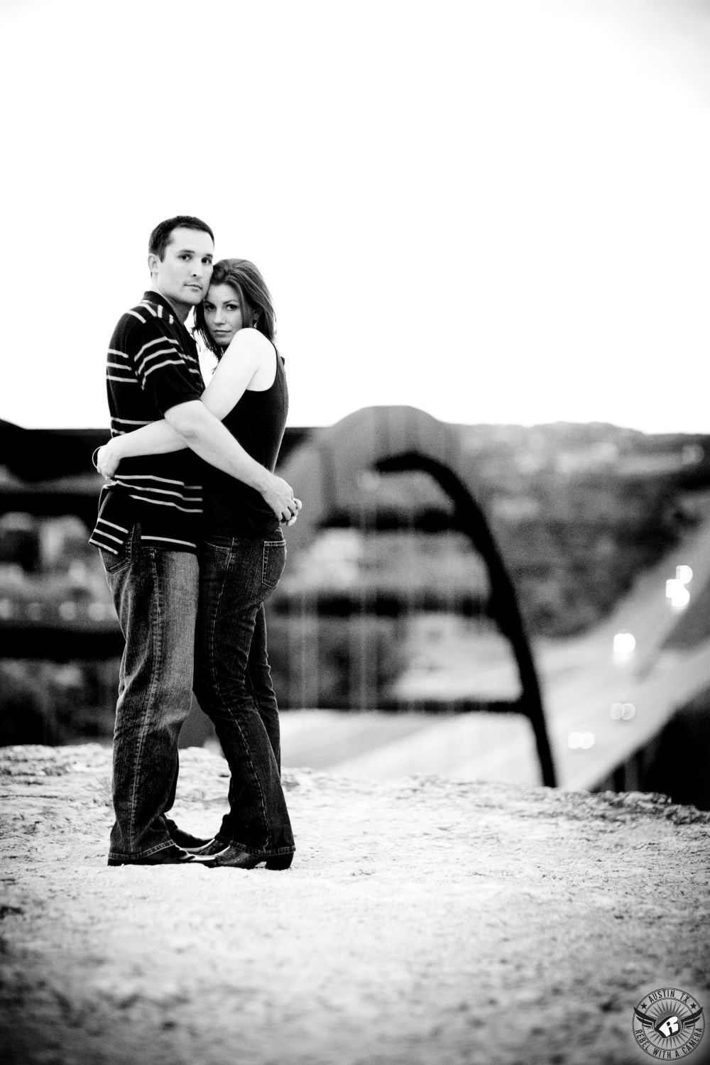 Sultry brunette wearing a black tank top, dark jeans and black books embraces guy with dark hair hearing a striped collared shirt and faded blue jeans on the cliff overlooking the 360 Bridge on Lake Austin in this black and white breathtaking engagement in Austin. 
